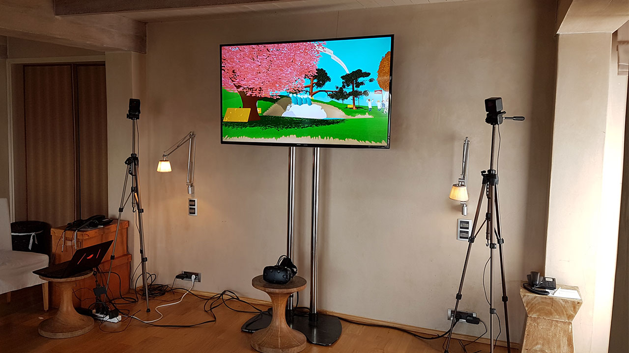 An indoor VR setup showing a television screen with a bright, cartoon-style VR environment. The space is equipped with VR headsets and microphones, prepared for Takeda employees to engage with the experience.