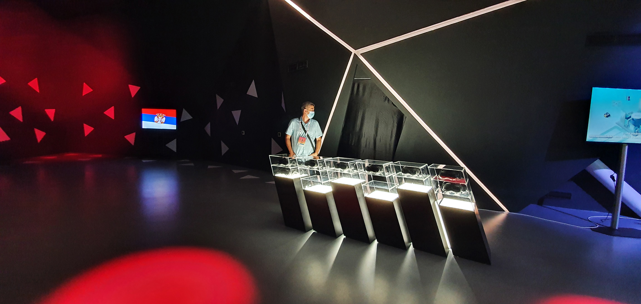 A single user engaging with a VR headset at the Serbian Pavilion, standing at a station designed with futuristic aesthetics. The area is lit by dramatic accent lighting that casts geometric shapes on the walls, emphasizing the Pavilion's focus on combining technology with cultural heritage.